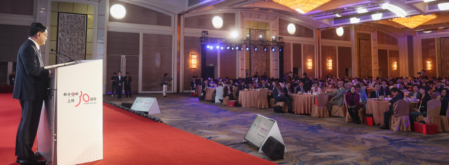 Speaker at podium in large event hall with seated audience, stage lights, and red carpet.